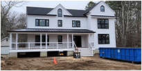 A large, white, two-story house under construction with black-framed windows, a front porch, and nearby blue dumpster. The setting feels calm and unfinished.