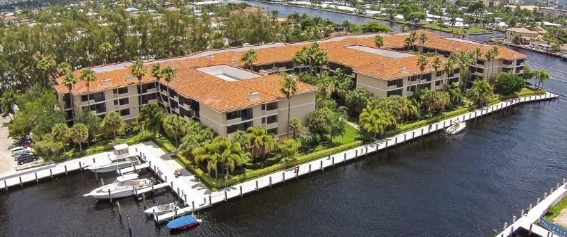 Aerial view of a waterfront apartment complex with terracotta roofs and lush green landscaping. A serene canal borders one side, adding tranquility.