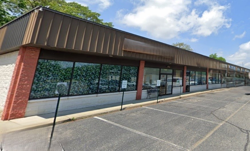 A closed storefront with large, reflective windows and brown overhead awning. Empty parking lot in front, under a partly cloudy blue sky. Quiet atmosphere.