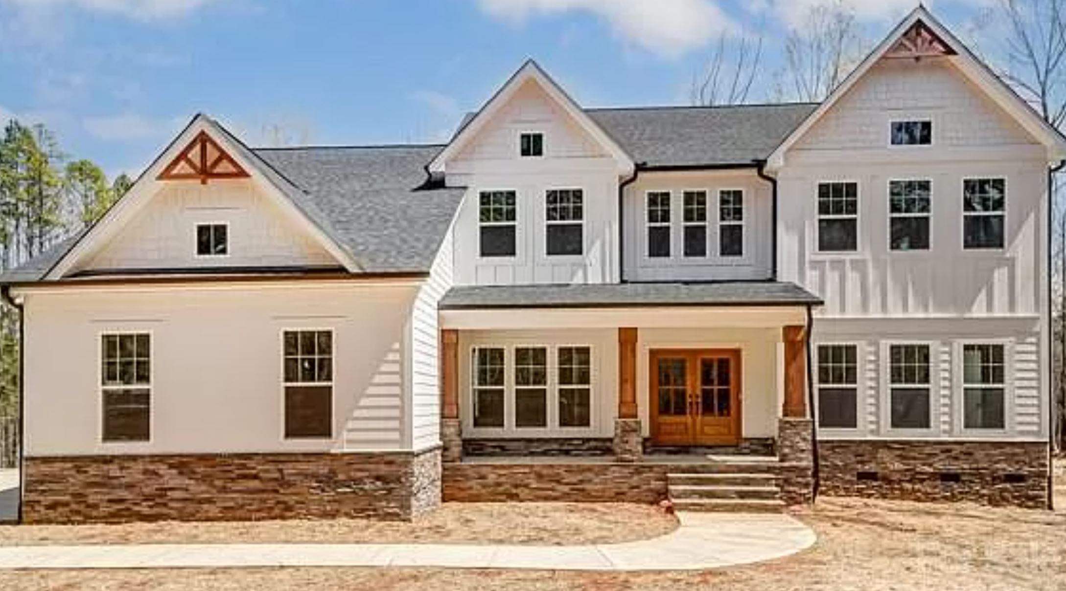 A modern two-story house with white siding, stone accents, and a dark roof. Features a welcoming front porch with wooden doors and large windows under a blue sky.