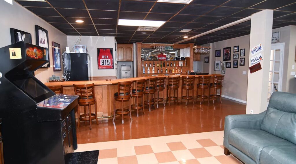 A cozy home bar with wooden stools, a countertop, and shelves stocked with bottles. The floor has a checkered pattern, and sports memorabilia decorates the walls.