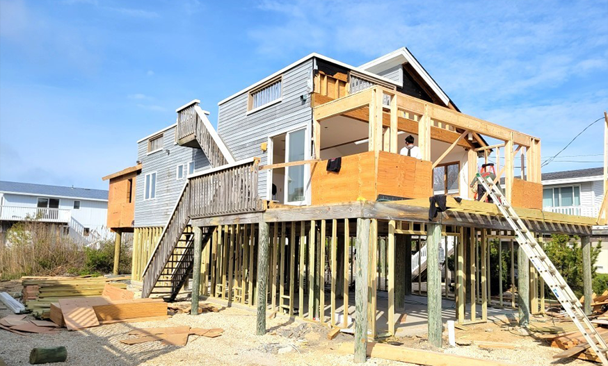 Elevated beach house under construction, featuring wooden beams, open framework, and stairs. Clear blue sky and a sandy ground create a coastal vibe.