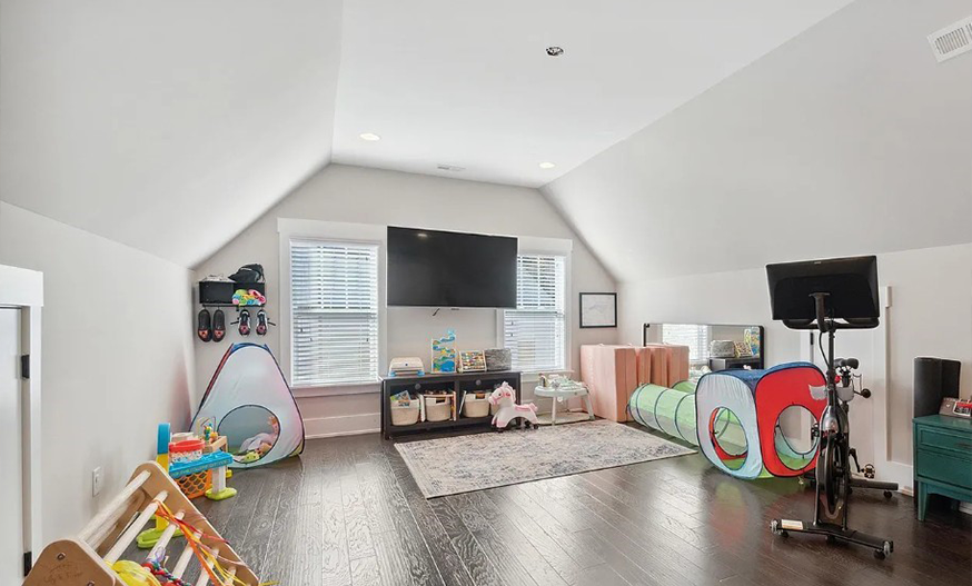 Spacious, bright playroom with a pitched ceiling, wooden floor, and two large windows. Toys, a play tunnel, and kid-friendly decor create a cheerful atmosphere.
