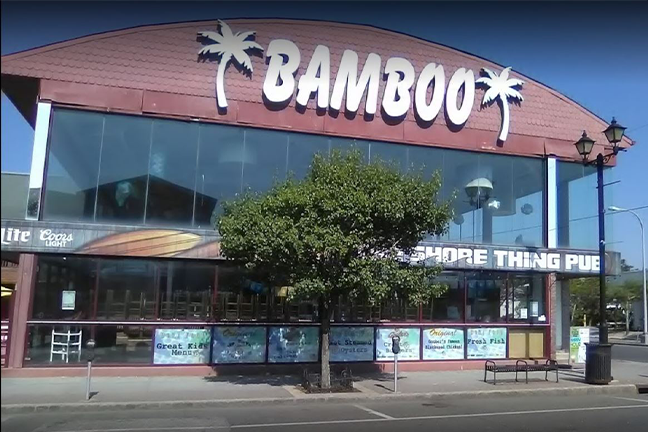 Front view of a tropical-themed building named "Bamboo" with large glass windows and palm tree decorations, set against a clear blue sky. A tree is in front.