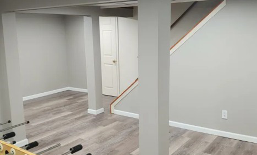 A modern basement with gray walls, light wood floors, and supporting columns. A closed white door is in the background, creating a clean, minimalist look.