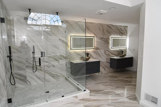 Spacious modern bathroom with glass shower, marble tile walls, sleek vanities with illuminated mirrors, and a large window, creating a luxurious feel.