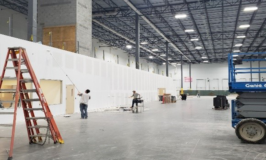A spacious warehouse interior with high ceilings and workers painting a long white wall. The space feels industrious, open, and in progress.
