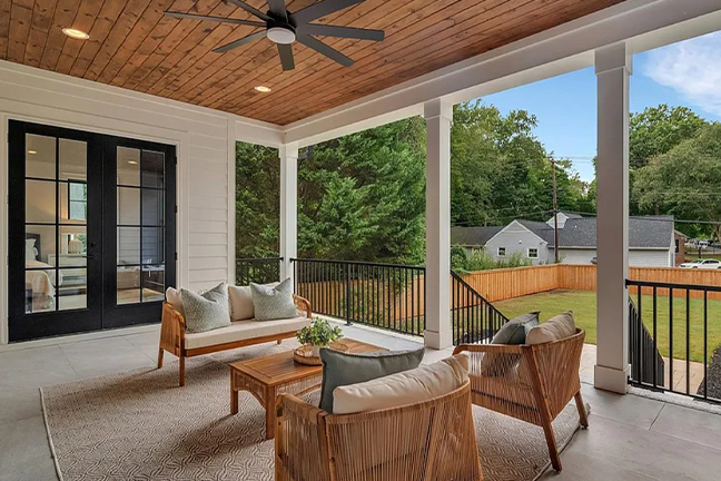 Chic outdoor patio with wooden ceiling, modern ceiling fan, wicker furniture, and plush cushions, overlooking a fenced green yard and trees. Cozy and inviting.