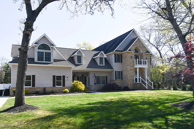 Large suburban house with gray siding, stone accents, and multiple gables. It features a well-maintained lawn, trees, and bright sunny skies.