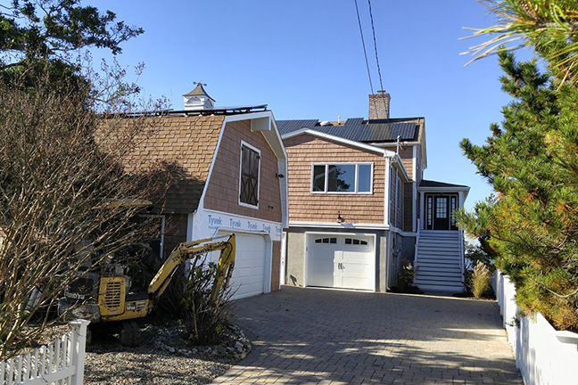 A two-story house with brown siding and white trim on a sunny day. An excavator is near the driveway, suggesting construction work. Blue sky overhead.