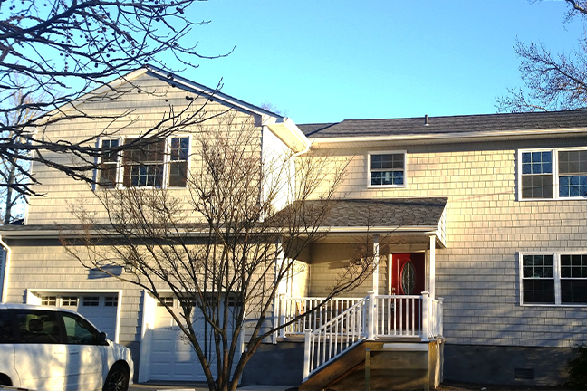 Two-story suburban house with beige siding, a red front door, and large windows. A bare tree stands in front, and the sky is clear and blue.