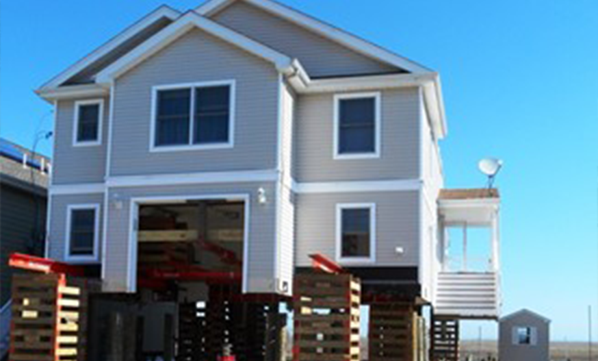 A raised beach house on stilts with a gray exterior, white trim, and large windows, set against a clear blue sky, conveying a coastal, airy feel.