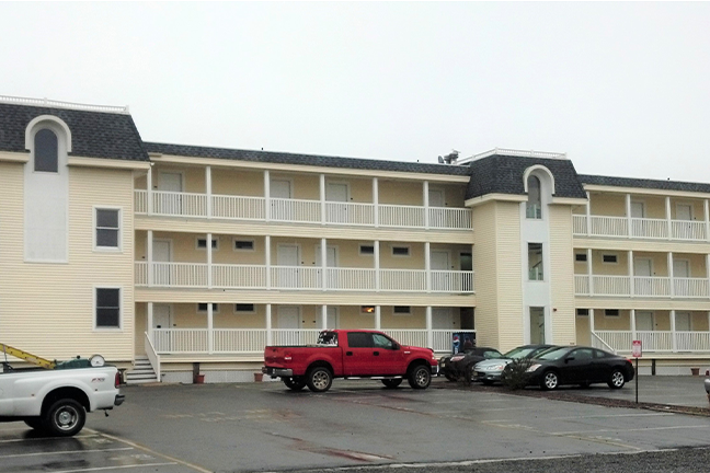 A three-story beige motel with balconies, a red truck, and cars in the parking lot under an overcast sky, conveying a quiet and ordinary atmosphere.