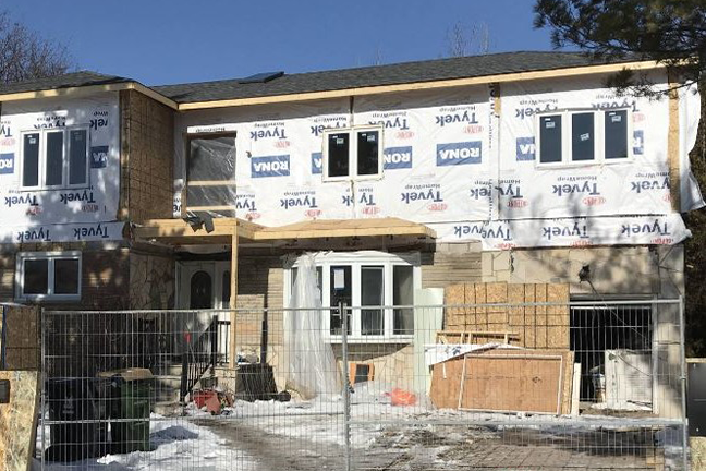 A two-story house under construction with wrapped walls, exposed wood, and unfinished windows. Snow covers the ground, and building materials are scattered around.