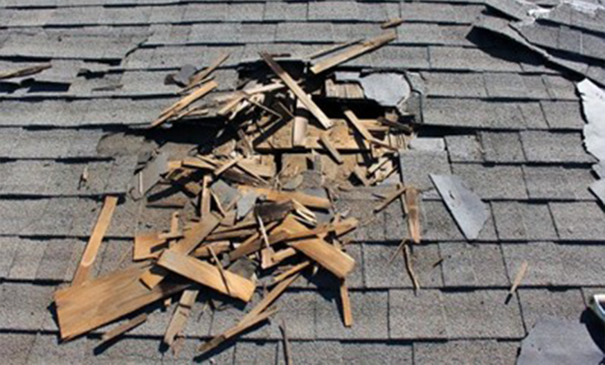 Damaged roof with a gaping hole surrounded by scattered wooden debris on gray shingles, conveying a sense of disrepair and vulnerability.