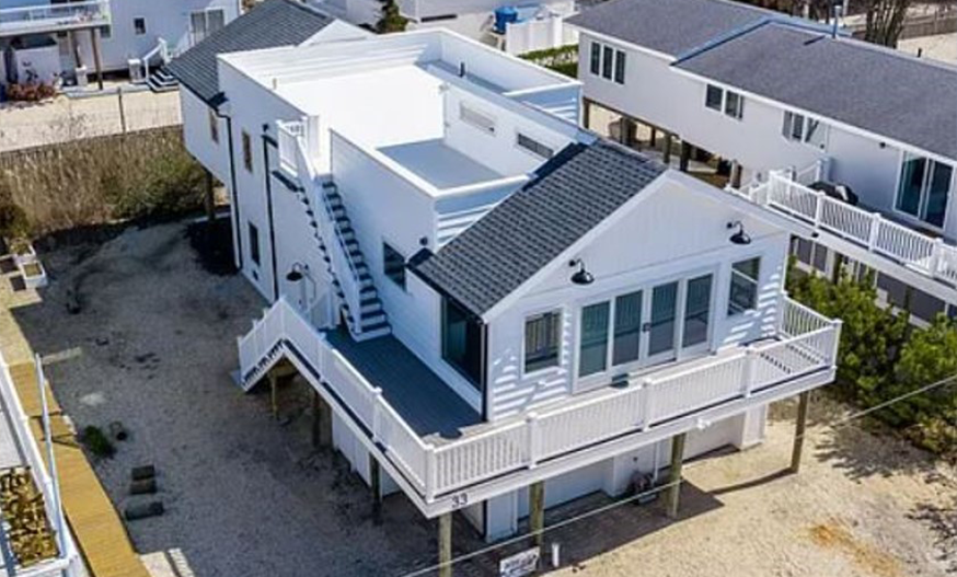 Aerial view of a modern, elevated white beach house with dark roof, surrounded by a sandy lot and neighboring homes, evoking a serene coastal vibe.