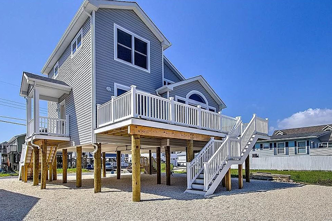 Elevated gray beach house on stilts with a white railing balcony and stairs, set against a clear blue sky, conveying a serene and coastal vibe.