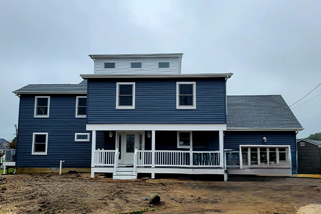 Blue two-story house with white trim and a covered porch under a cloudy sky. The sandy ground suggests ongoing construction or recent completion.