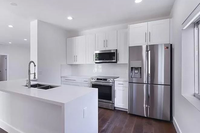 Modern kitchen with white cabinets, stainless steel appliances, and dark wood flooring. An island counter is in the foreground under bright lighting.