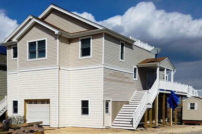 A two-story beige house with a slanted white roof, large windows, and an elevated white staircase against a bright blue sky with fluffy clouds.
