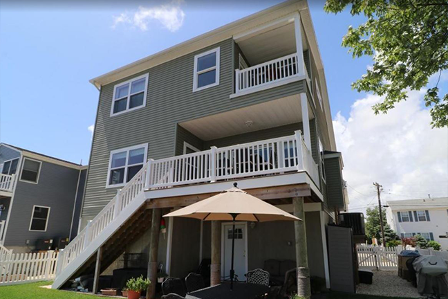 Three-story house with green siding and white railings, featuring balconies and a patio with a sun umbrella. Bright, sunny day with blue skies.