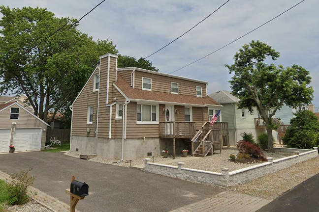 A two-story house with brown siding and a front porch, featuring stairs and an American flag. A driveway and mailbox are in the foreground, trees in the background.