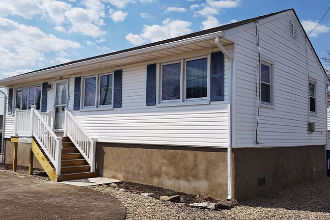 A white single-story house with multiple windows, set against a blue sky with clouds. A small staircase leads to the entrance. Calm and inviting atmosphere.