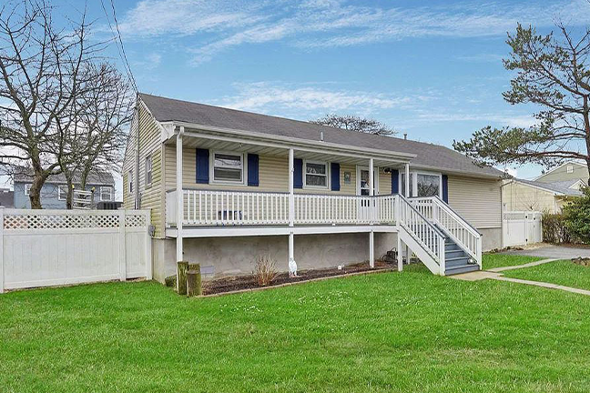 A beige single-story house with a long front porch, blue shutters, and white railing. It sits on a lush green lawn under a partly cloudy blue sky.