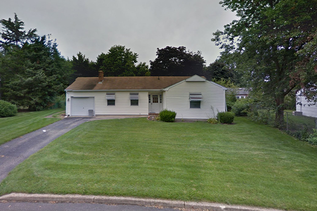 Single-story white house with attached garage, large front lawn, and hedges. Overcast sky and trees in the background, conveying a calm, suburban feel.