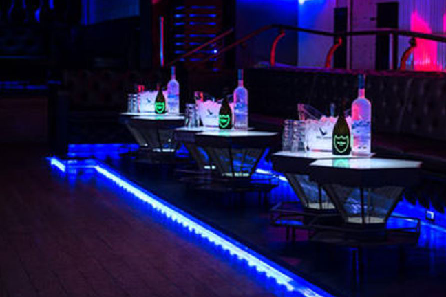Dimly lit nightclub setting with tables holding bottles of water, ice buckets, and glasses. Neon blue lights create a lively, modern ambiance.