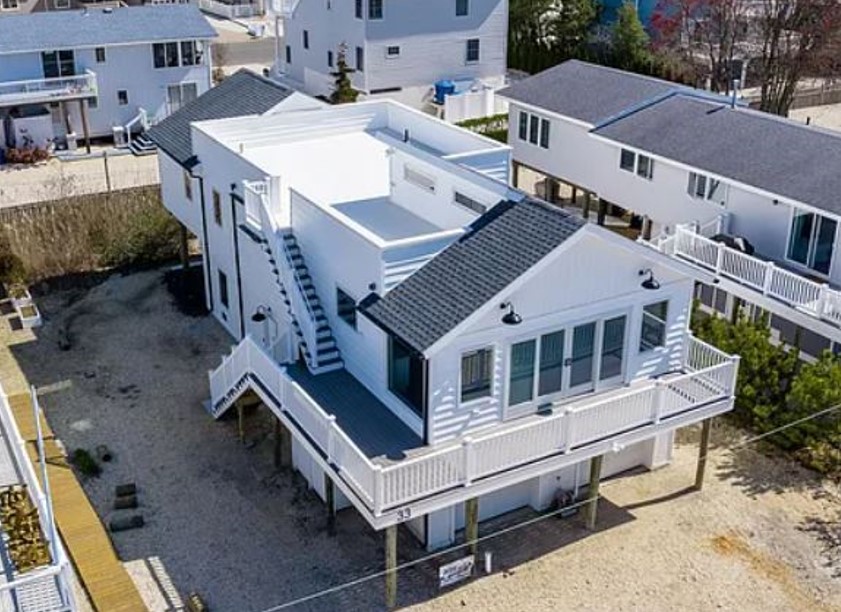 Aerial view of a modern white beach house on stilts, featuring large windows, multiple balconies, and an exterior staircase, surrounded by similar homes.