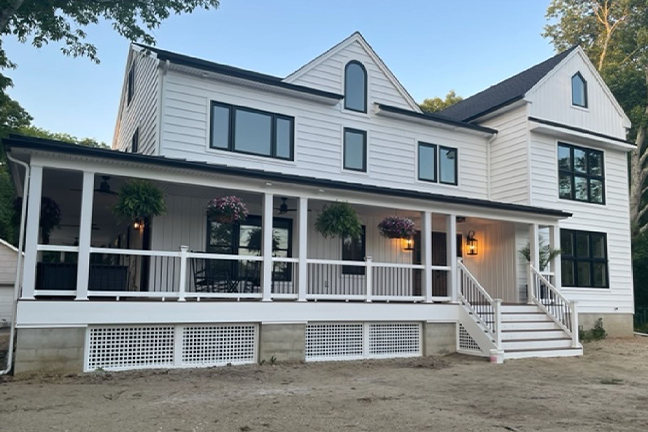Two-story white house with black-trimmed windows, a covered porch, hanging plants, and warm exterior lights. A calm, welcoming atmosphere is conveyed.