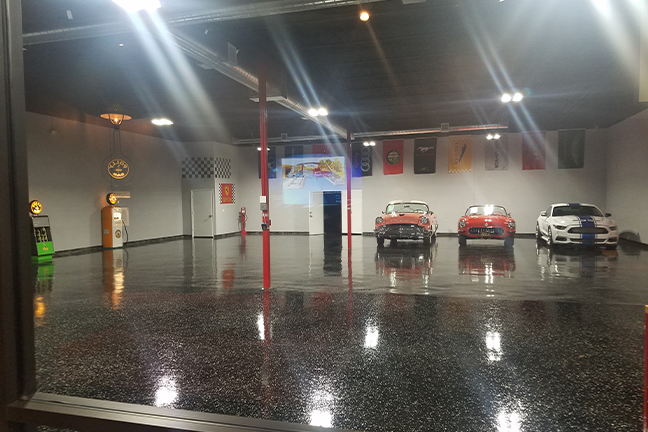 A polished garage with two red vintage cars parked in front of a white wall adorned with flags. Bright ceiling lights create reflections on the shiny floor.