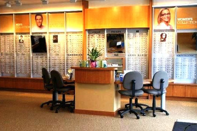 Optometrist office with a display of glasses on well-lit walls. A wooden desk with chairs and a plant conveys a welcoming and professional atmosphere.