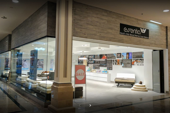 A modern mattress store with large glass windows displays various beds inside. A sale sign is visible, creating a welcoming and inviting atmosphere.