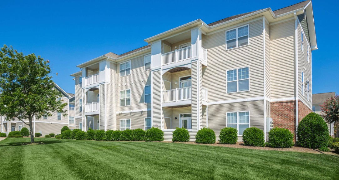 A three-story apartment building with beige siding and white balconies, set against a clear blue sky. Well-kept lawn and small green bushes line the front.