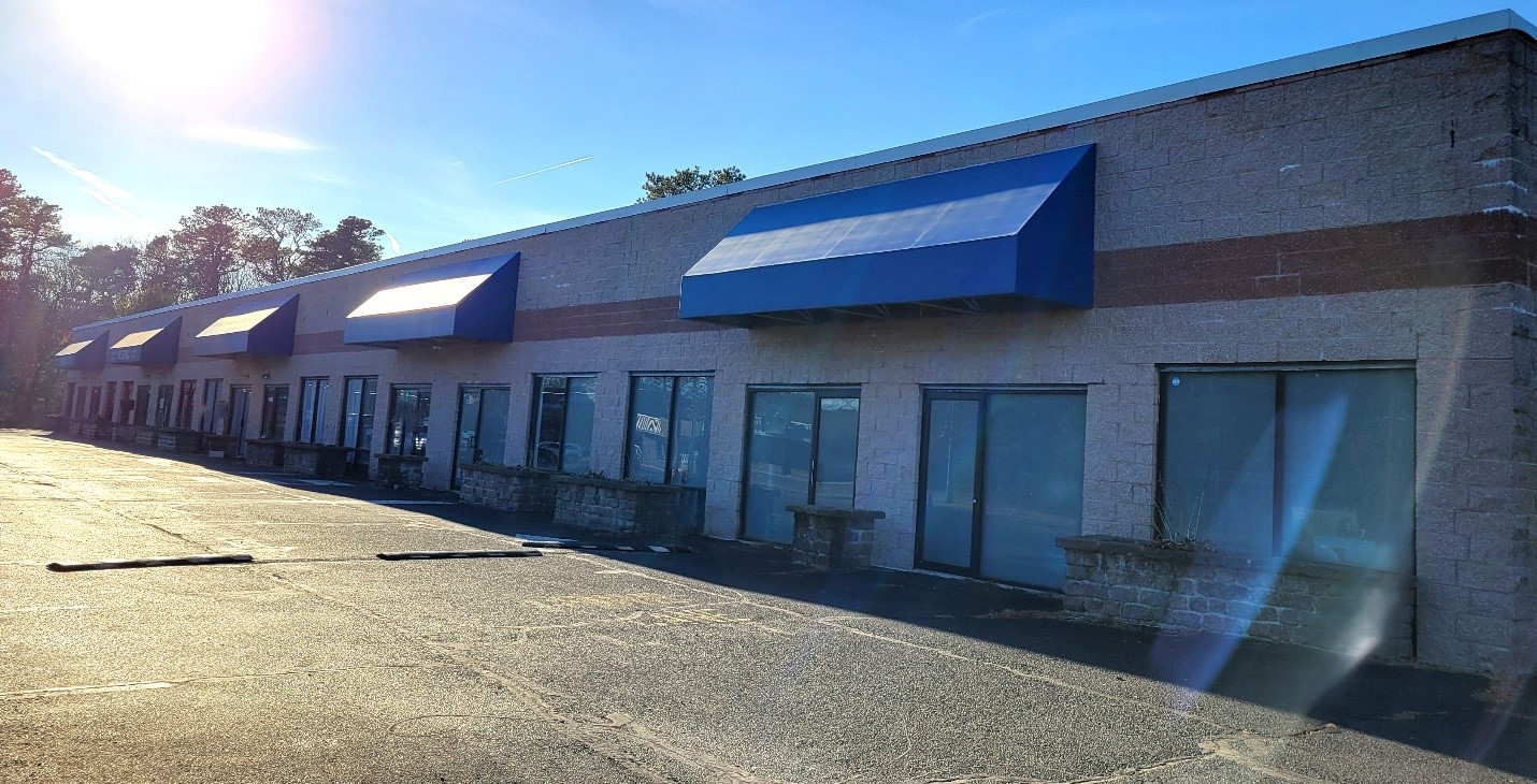A row of closed storefronts with blue awnings and large windows under a clear blue sky. The empty parking lot suggests inactivity and solitude.