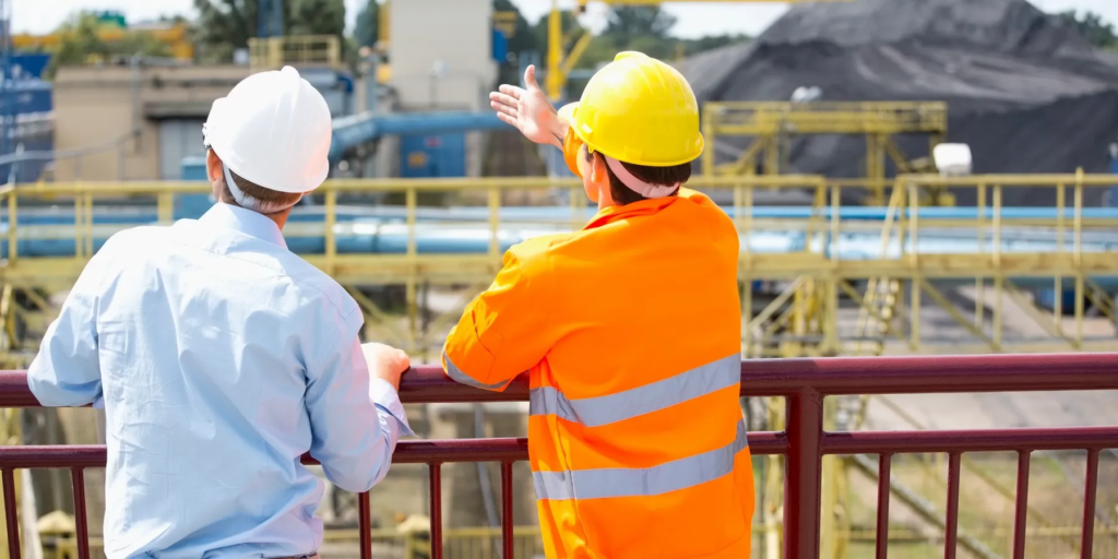 Two construction workers in safety gear, one in an orange vest and yellow helmet, facing industrial equipment, discussing the site with a focused stance.