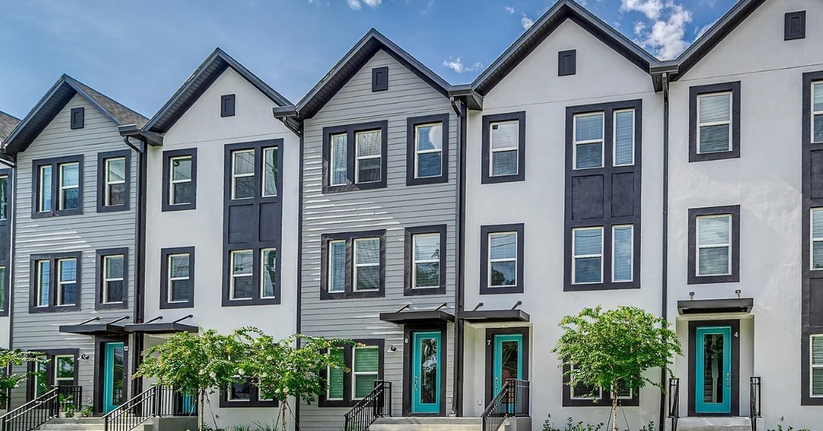 Row of modern townhouses with gray and white siding, blue front doors, and tall windows. Small trees line the street, under a clear blue sky.