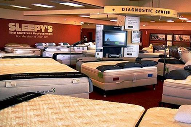 Showroom filled with neatly arranged mattresses on display under bright lighting. A diagnostic center sign and a screen are visible, creating a welcoming retail atmosphere.