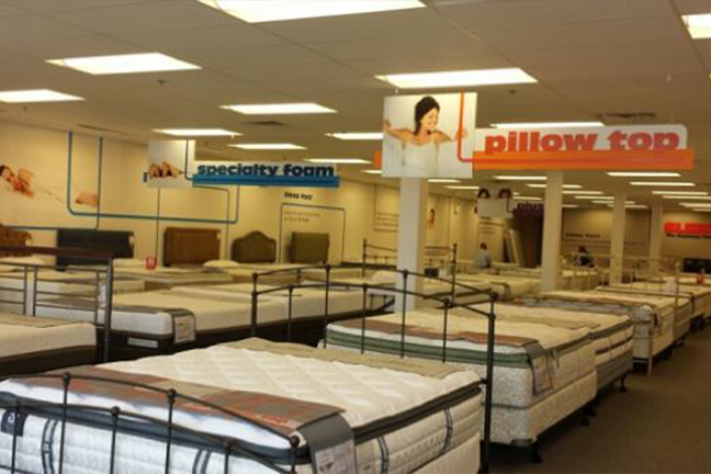 Mattress store interior with neatly arranged beds and promotional signs overhead for quality and pillows. Bright lighting creates a welcoming atmosphere.
