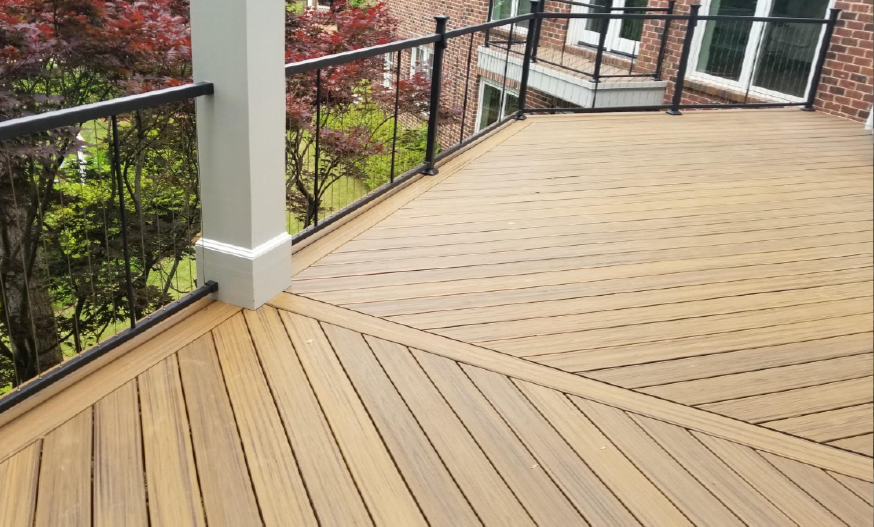 A spacious wooden deck features a geometric pattern and black railing, with lush greenery and a brick building visible in the background.