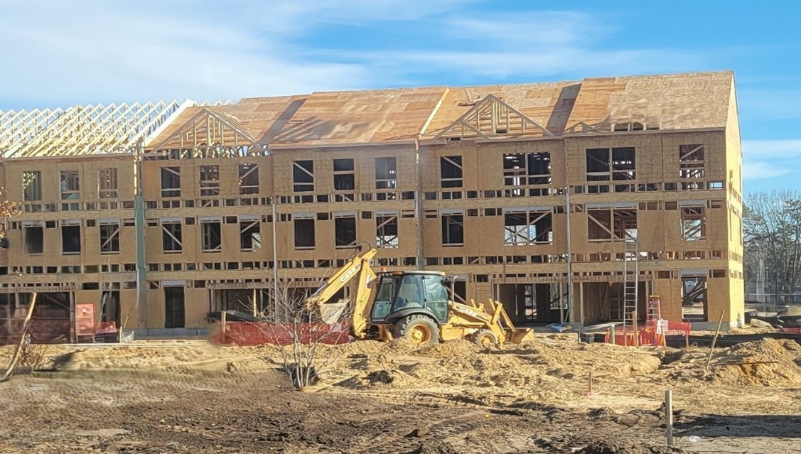 Construction site of a large building with wooden framework, scaffolding, and a yellow bulldozer in front. The sky is clear and blue.