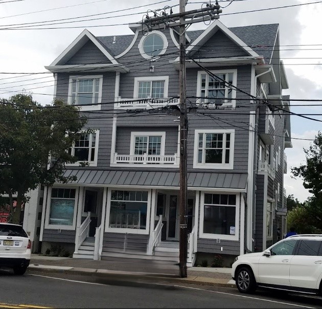 Three-story gray building with white trim and multiple windows, located on a street with parked cars. Overhead power lines and trees are visible.