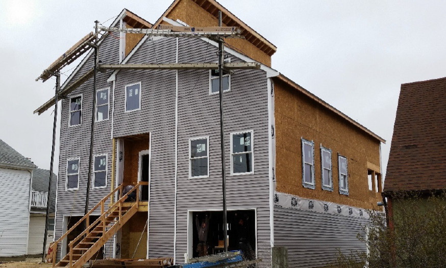 A two-story house under construction with an unfinished wooden facade and exposed insulation. Cloudy sky in the background adds to the work-in-progress feel.