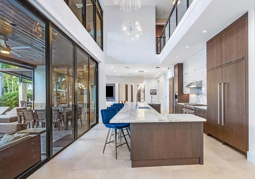 Modern kitchen with high ceilings and glass walls, showcasing a long marble island with blue chairs. Bright, spacious, and elegant atmosphere.
