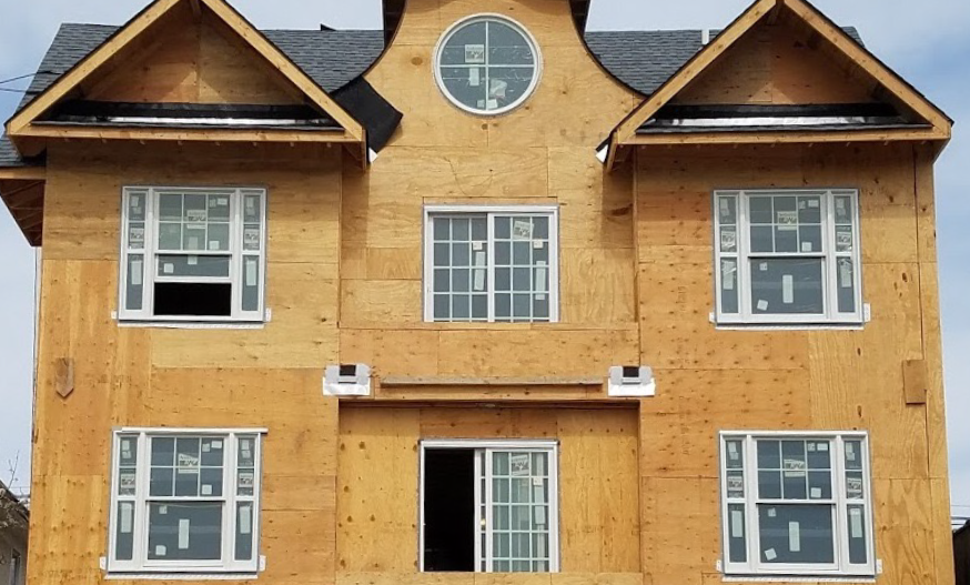 A multi-story house under construction with exposed wood walls and installed windows. The roof has a round window and steep gables, conveying a sense of progress.