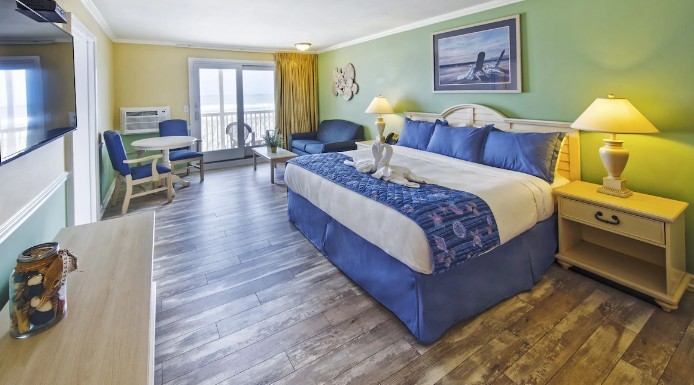 Spacious hotel room with a king-sized bed in blue and white linens, wooden floor, sea-themed decor, and a large window overlooking a balcony.