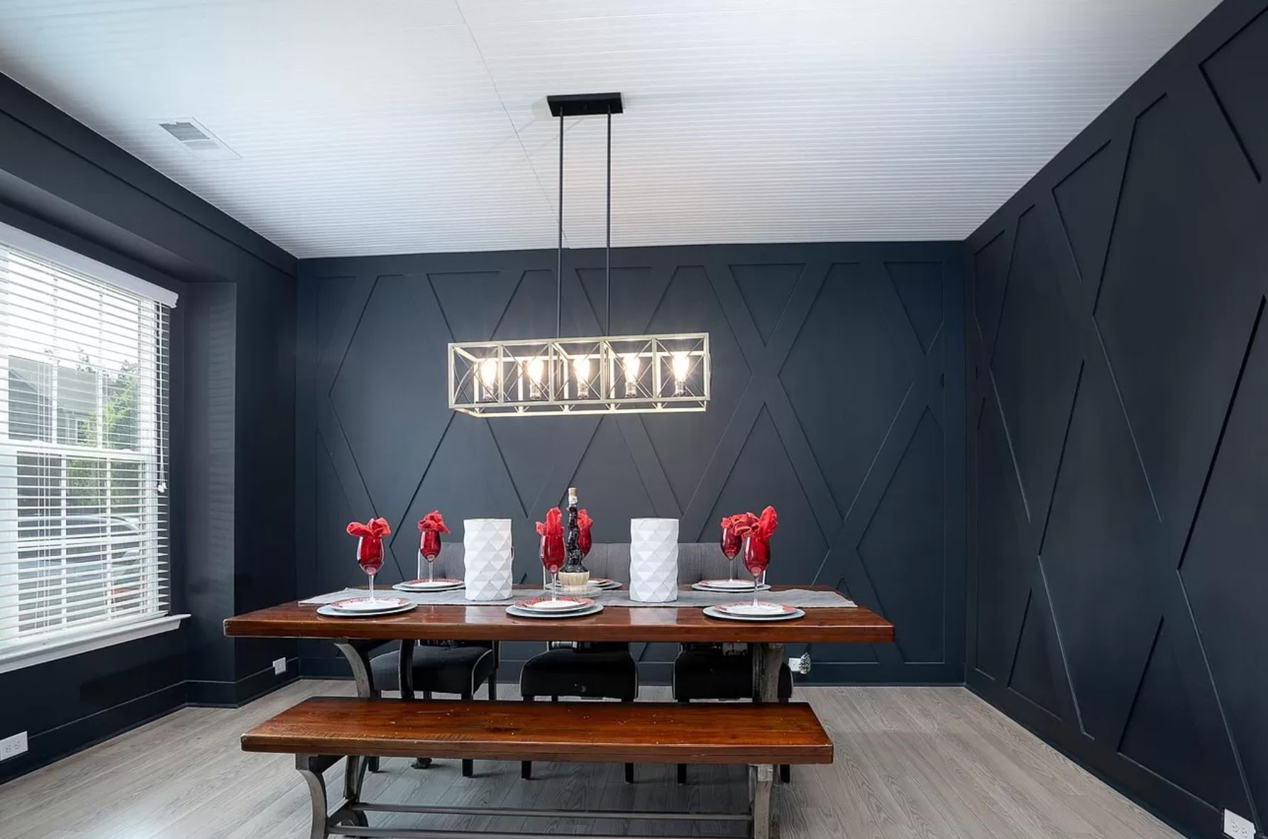 Modern dining room with dark walls featuring a wooden table set for six. Red flowers and white candles decorate the table. A sleek chandelier hangs above.