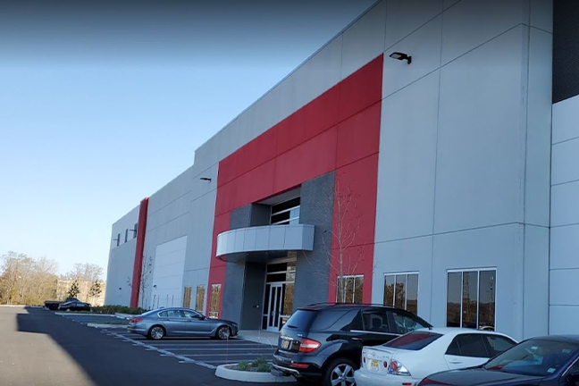 A modern industrial building with a red and gray facade, large rectangular windows, and a metal awning. Several cars are parked outside under a clear blue sky.